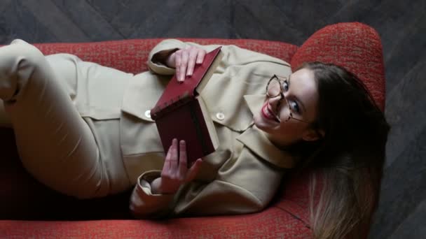 Bella bionda giovane donna in occhiali e abito beige seduta sul divano rosso durante la lettura di libri e guardando giocosamente nella fotocamera. Vista dall'alto — Video Stock