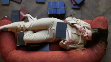 Kırmızı kanepede gözlüklü çekici rüya gibi kız sevgi mektubu okuyor ve neşeyle yerde birçok kitap la çevrili gülümsüyor