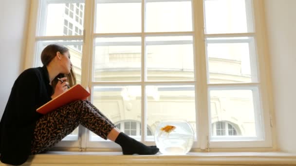 Hermosa joven soñadora escribiendo en el cuaderno y posando en el alféizar de la ventana con acuario con peces dorados en él — Vídeos de Stock