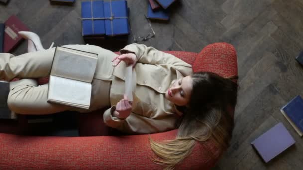 Atractiva chica de ensueño en gafas en el sofá rojo leyendo carta de amor y sonriendo alegremente rodeado de muchos libros en el suelo — Vídeo de stock