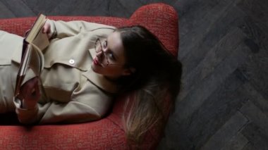Kitap okurken ve eğlenceli bir şekilde el çağırırken kırmızı kanepede oturan bej takım elbiseli gözlüklü çekici genç kadın. Üstte görüntü
