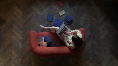 Duygusal genç kadın manifatura modern laptop kanepe ve mutlu bir şekilde evde katta çok kitap tarafından çevrili gülüyor. Üstten Görünüm