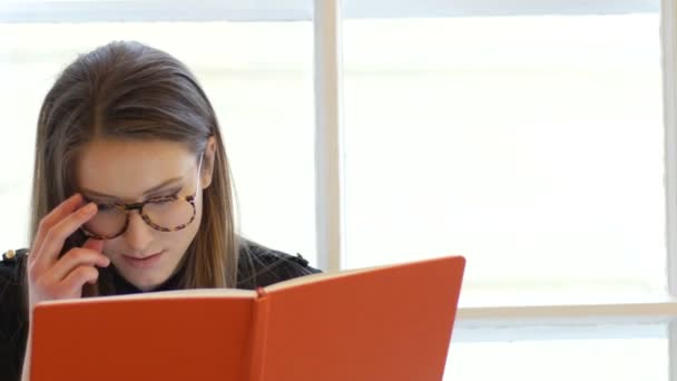Retrato de una joven estudiante escribiendo en la libreta con gafas sentadas en el alféizar de la ventana — Vídeo de stock