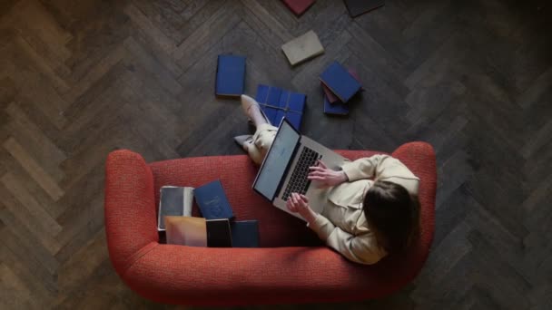 Känslomässiga ung kvinna textning på modern laptop på soffa och lyckligt skrattar omgiven av många böcker på golvet hemma. Ovanifrån — Stockvideo