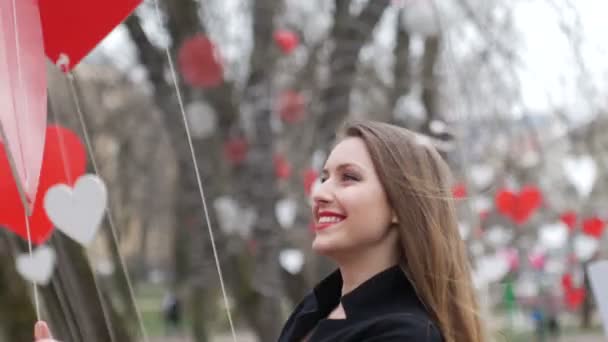 Elegante chica rubia sonriendo alegremente y jugando con la decoración de corazones de papel rojo en el parque de otoño. Celebración de San Valentín — Vídeos de Stock