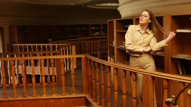 En una cálida luz pálida en la vieja biblioteca hermosa mujer joven con traje y gafas elegir un libro para leer — Vídeos de Stock