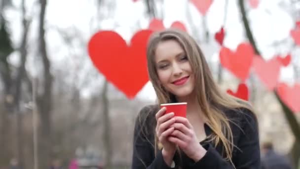 Νεαρή όμορφη ξανθιά hipster γυναίκα ποζάρουν στο πάρκο φθινόπωρο διακοσμημένα με κόκκινο χαρτί καρδιές φόντο με φλιτζάνι καφέ takeaway. Ημέρα του Αγίου Βαλεντίνου έννοια — Αρχείο Βίντεο