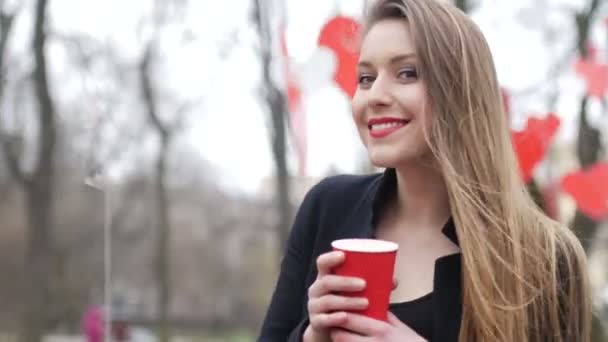 Jovem mulher hipster loira bonita posando no parque de outono decorado com fundo corações de papel vermelho com xícara de café takeaway. Conceito do Dia dos Namorados — Vídeo de Stock