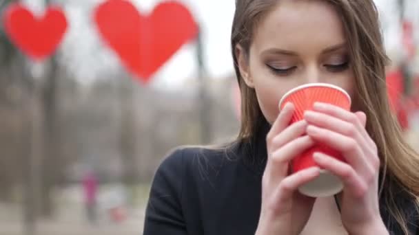 Молодая красивая блондинка хипстер женщина позирует на осеннем парке украшены красной бумаги сердца фона пить чашку кофе на вынос. День святого Валентина — стоковое видео