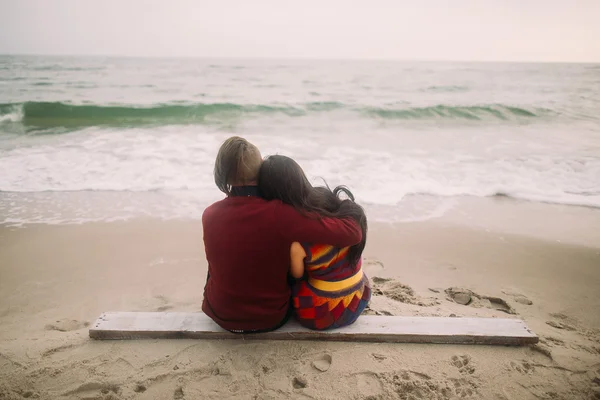Pretty young couple sitting on the beach and embracing. Winter sea background. Tender moment. Back view — Stock Photo, Image