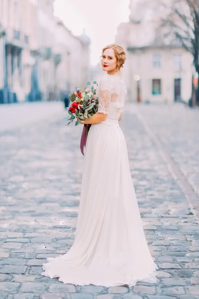 Charmante Braut im langen Spitzenkleid mit Vintage-Bouquet, das über die Schulter in die Kamera schaut, mit alter Stadtarchitektur im Hintergrund — Stockfoto