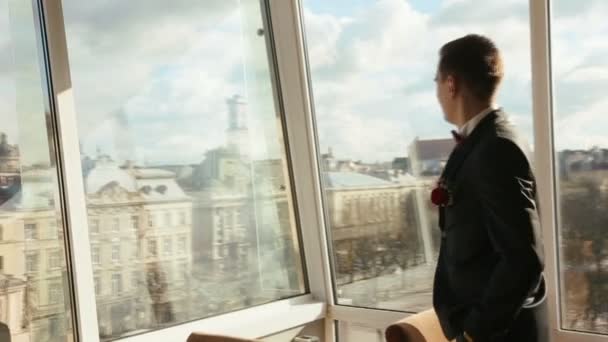Красивий впевнений чоловік у чорному костюмі стоїть біля скляної стіни в кафе. Дивовижний міський пейзаж Львова на фоні — стокове відео