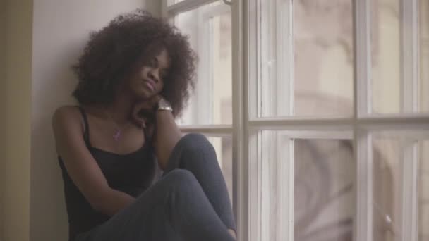 Beautiful and sad young african woman sitting on the windowsill and grieving — Stock Video