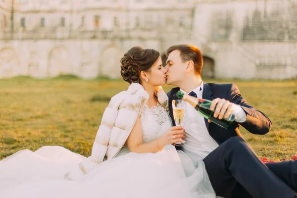 Young bride and groom sitting on picnic, drinking champagne   from glasses at wedding — Zdjęcie stockowe