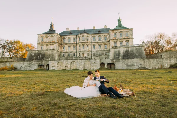 Schöne Braut und Bräutigam sitzt auf Decke kariert mit Champagner in der Nähe der alten Villa bei Sonnenuntergang — Stockfoto