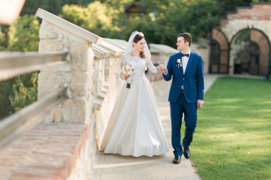 Young bride in white dress and groom walking holding hands near old castle