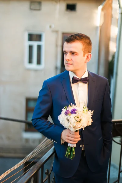 Mignon marié élégant en costume bleu et noeud papillon avec un bouquet de roses debout au balcon regardant vers — Photo