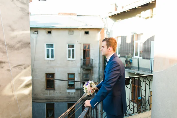 Mignon marié élégant en costume bleu et noeud papillon avec un bouquet de roses debout au balcon regardant vers — Photo