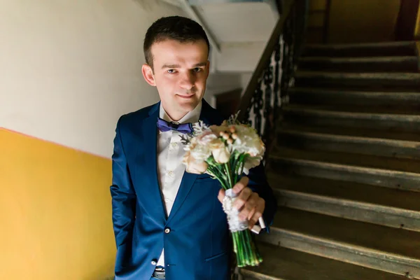 Elegante noivo elegante no terno azul e arco com buquê de rosas escadas de fundo — Fotografia de Stock