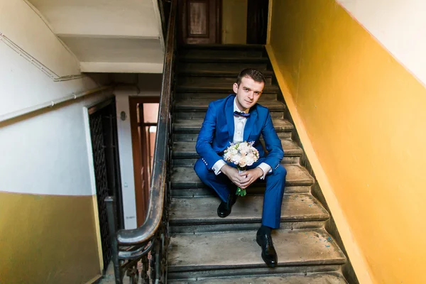 Jovem atraente sorridente homem de terno azul sentado em escadas com buquê, tiro em casa — Fotografia de Stock