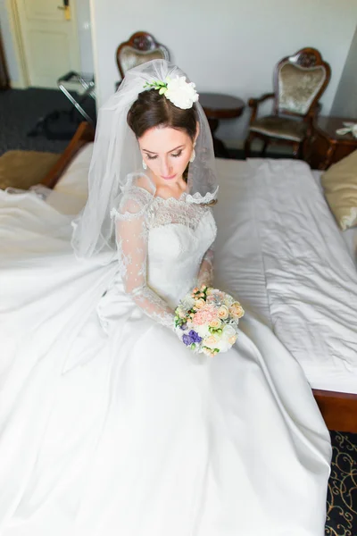 Linda noiva jovem com maquiagem, penteado extravagante em vestido branco e véu sentado na cama olhando para baixo — Fotografia de Stock