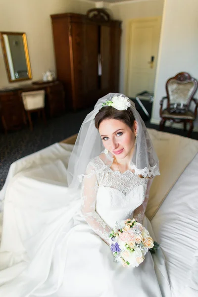 Linda noiva jovem com maquiagem, penteado extravagante em vestido branco e véu sentado na cama — Fotografia de Stock