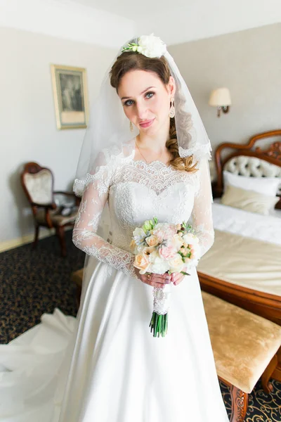 Incrivelmente bela, jovem noiva posando no quarto de hotel em um vestido magnífico e delicado buquê — Fotografia de Stock