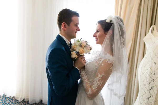 Hermosa pareja de boda amorosa está sosteniendo ramo de flores y besos en el fondo interior del hotel de lujo —  Fotos de Stock