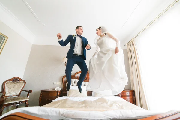 Beautiful happy bride and groom jumping on the bed in rich hotel interior