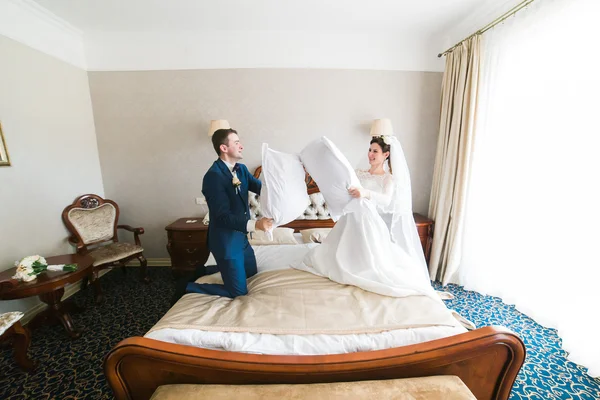 Otel odasında yatakta yastıklarla kavga eden mutlu gülümseyen yeni evli çiftin portresi — Stok fotoğraf