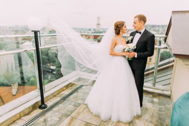 Mutlu gelin ve damat yumuşak cityscape arka plan ile terasta sarılma, rüzgar uzun gelin peçe kaldırma