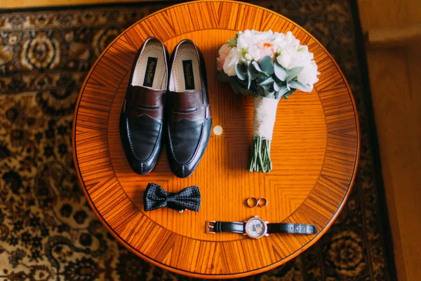 Acessórios masculinos: sapatos pretos e gravata borboleta, relógio, anéis na mesa de madeira marrom. Buquê de noiva — Fotografia de Stock