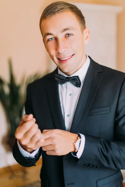 Elégant jeune homme de mode en smoking noir ajustant ses boutons de manchette tout en regardant la caméra à l'intérieur — Photo