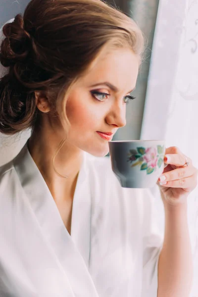 Portret van de close-up van de mooie bruid poseren in gewaad en drinken uit de beker in de buurt van venster op de ochtend van de bruiloft — Stockfoto