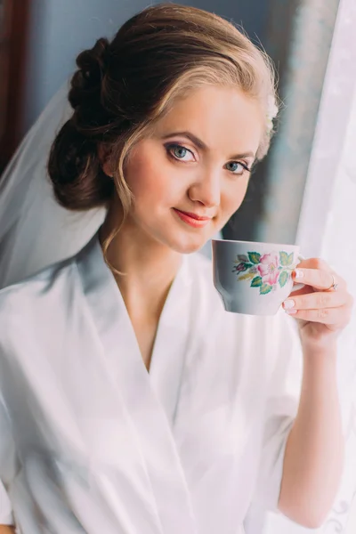 Portret van de close-up van de mooie bruid poseren in gewaad en drinken uit de beker in de buurt van venster op de ochtend van de bruiloft — Stockfoto