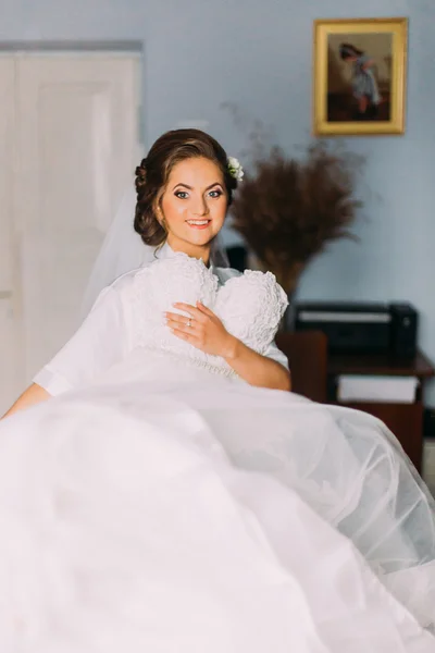 Beautiful sensual blonde bride in robe and white veil posing with wedding dress indoors — Stock Photo, Image