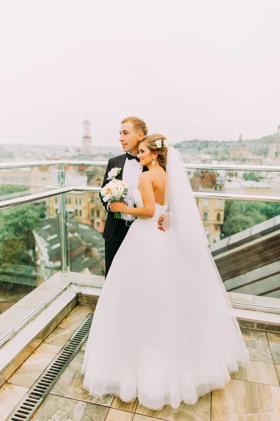 Счастливая невеста и жених мягко обнимаются на террасе с городским пейзажем — стоковое фото