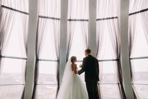 आधुनिक इमारत में आंतरिक पृष्ठभूमि पर सुंदर खिड़की पर हाथ पकड़ते हुए स्टाइलिश लक्जरी दुल्हन और दुल्हन — स्टॉक फ़ोटो, इमेज