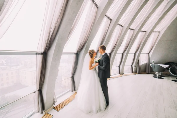 Красивая супружеская пара обращена лицом друг к другу и обнимается в белой комнате с большими окнами — стоковое фото