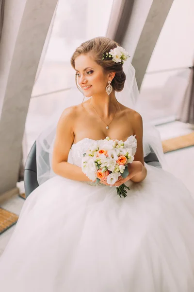 Close-up retrato de jovem noiva bonita em vestido branco e véu sentado perto da janela segurando buquê — Fotografia de Stock