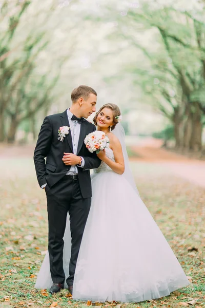 Full portrait of happy young wedding couple standing arm-in-arm in park — Stock fotografie