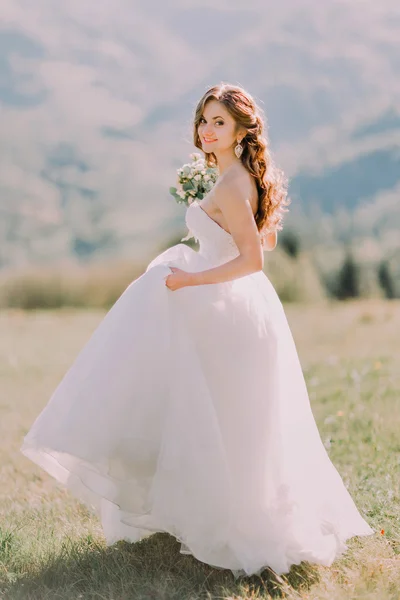 Beautiful blonde bride in  wedding dress runs across the field toward mountains — Stockfoto