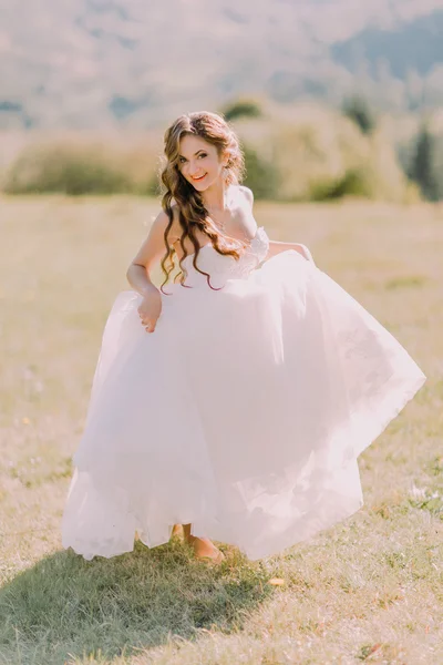 Beautiful blonde bride in  wedding dress runs across the field toward mountains — Stockfoto