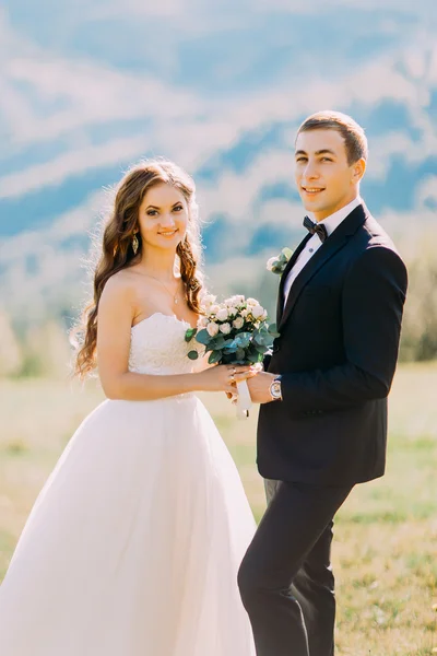 Όμορφη υπέροχη ευτυχισμένη νύφη με μπουκέτο και κομψό κουμπάκι κρατώντας τα χέρια στο φόντο των βράχων στα βουνά — Φωτογραφία Αρχείου