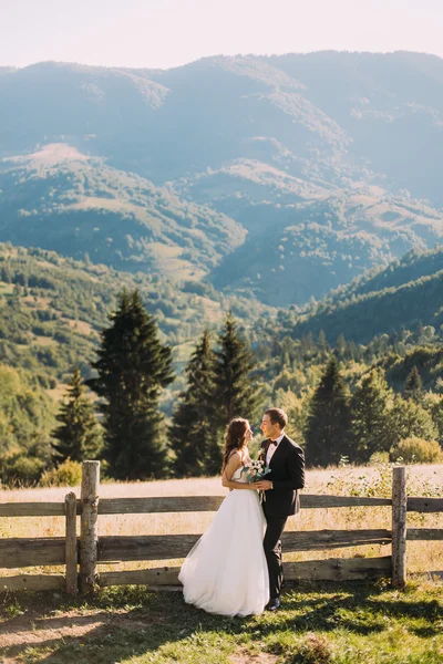 La novia y el novio de pie en el puente de madera en la naturaleza, abrazando cerca de valla con fondo de montaña —  Fotos de Stock