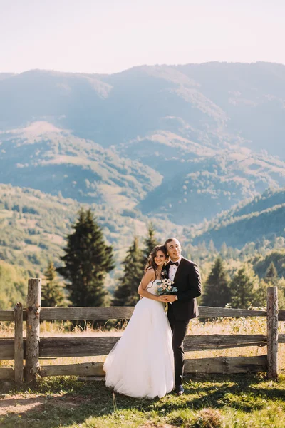 La novia y el novio de pie en el puente de madera en la naturaleza, abrazando cerca de valla con fondo de montaña —  Fotos de Stock