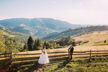 Beyaz elbiseli mutlu sarışın gelin ve damat ahşap çit yakınında ayrı duruyor