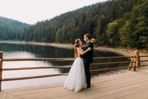 Luksusowe szczęśliwy wesele pary młodej bukiet i pana młodego, obejmując w pobliżu ogrodzenia na tle jeziora pod zachód słońca — Zdjęcie stockowe