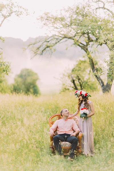 Mulher bonita vestindo vestido lilás e coroa segurando buquê de pé perto de homem bonito sentado na cadeira ao ar livre — Fotografia de Stock