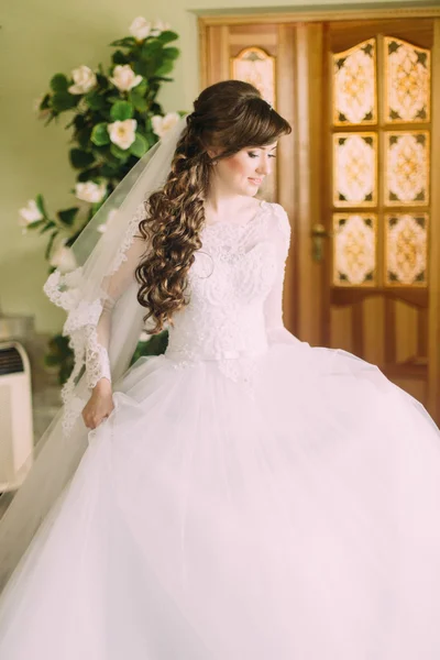 Hermosa novia en elegante vestido de novia blanco y velo con pelo largo y rizado posando en el interior —  Fotos de Stock